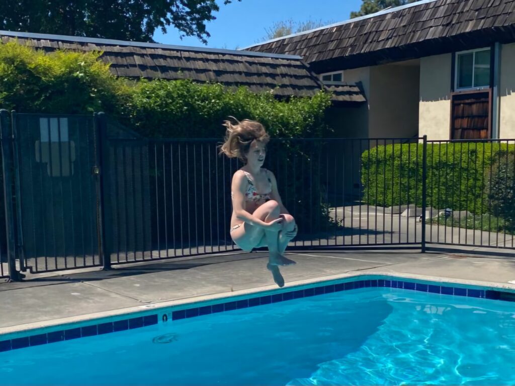 Person mid-jump performing a cannonball into a backyard swimming pool, knees tucked to chest and hair lifted by motion. A black metal fence, green hedges, and a house with a shingled roof are visible in the background under a clear blue sky.